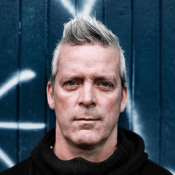 Close-up portrait of Sean Cranbury with spiky grey hair, wearing a black hoodie against a blue wooden wall with white graffiti.